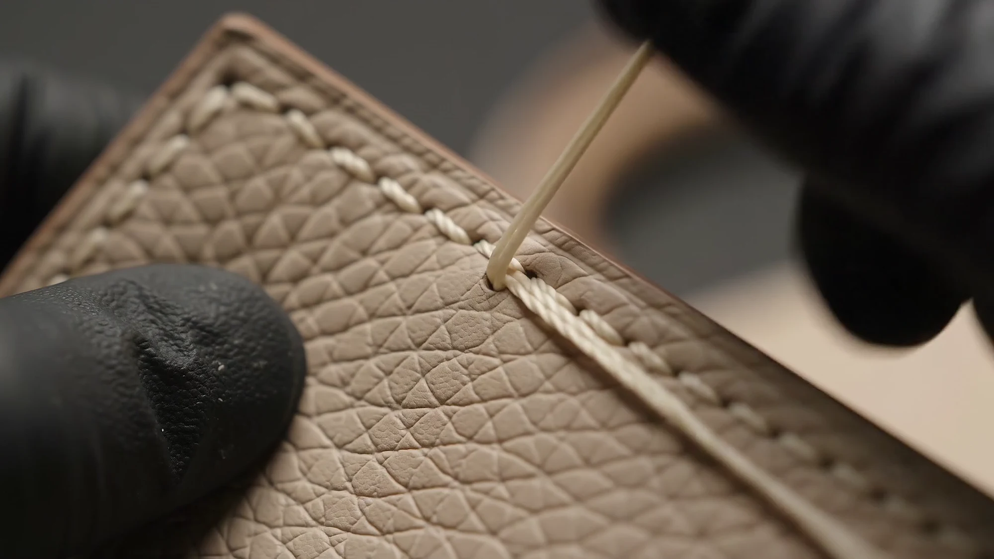 A needle passes through a line of pale stitches in grained leather