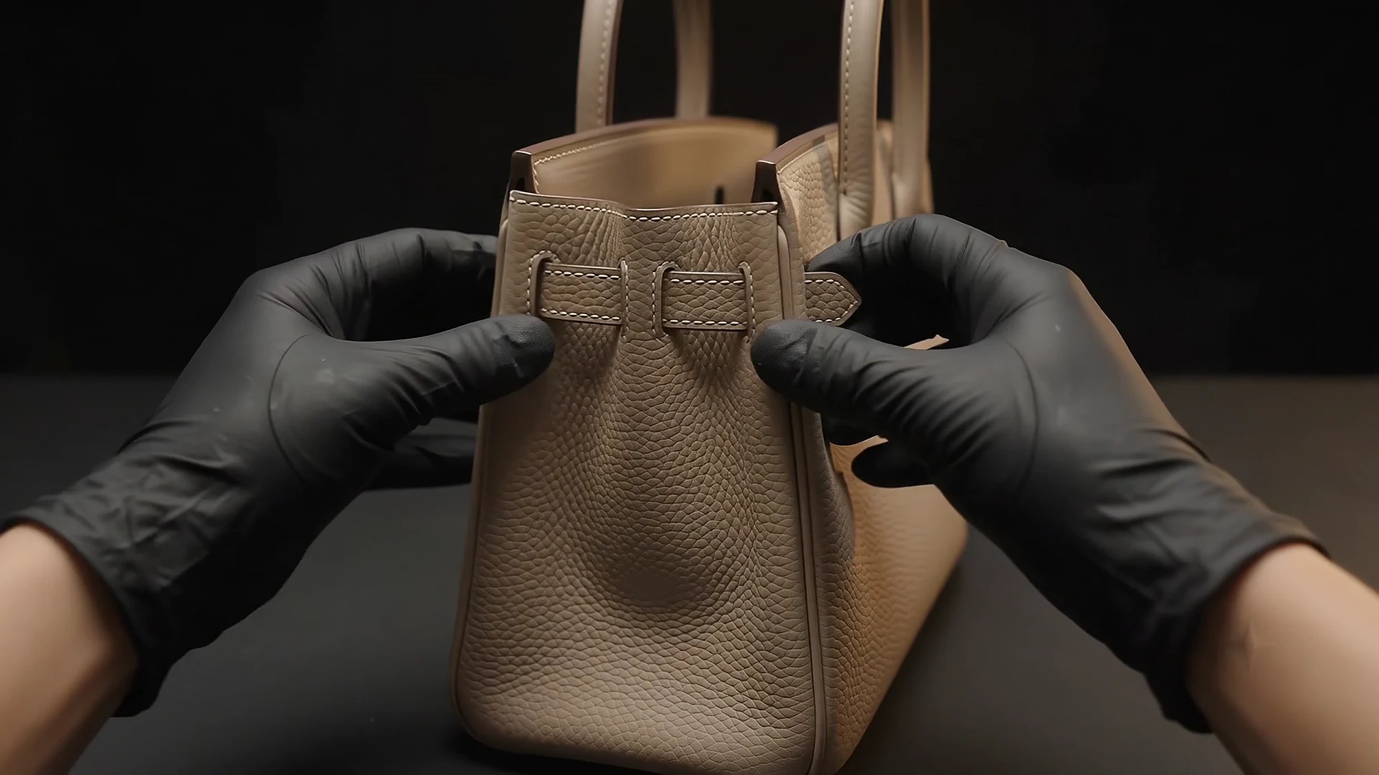 Two gloved hands hold the side panels of a bag together beside a row of fine stitches