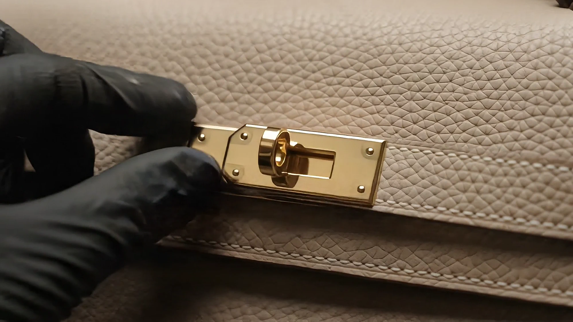 A gloved hand fits a gold lock to the flap of a taupe bag